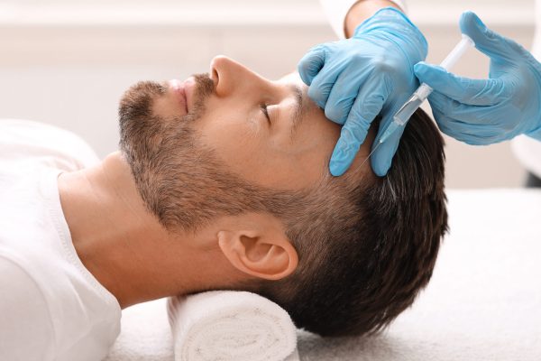 Side view of handsome middle aged man getting hair treatment at beauty salon. Man having mesotherapy session at aesthetic clinic, therapist hands in gloves making injection in scalp, closeup
